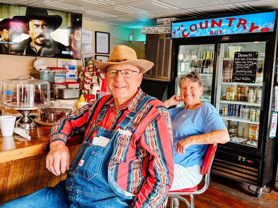 Two regulars at the counter — one in a straw hat and striped overalls — under the Country Boy cooler sign, the cake stand behind them.