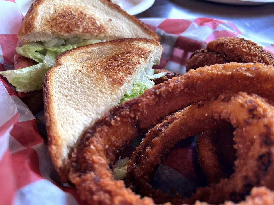 A BLT on Texas toast with thick hand-breaded onion rings in a red-checkered paper basket.