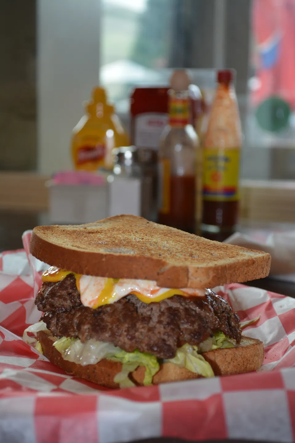 A double smash burger on toasted bread, up close in its checkered basket, the condiment bottles soft behind.