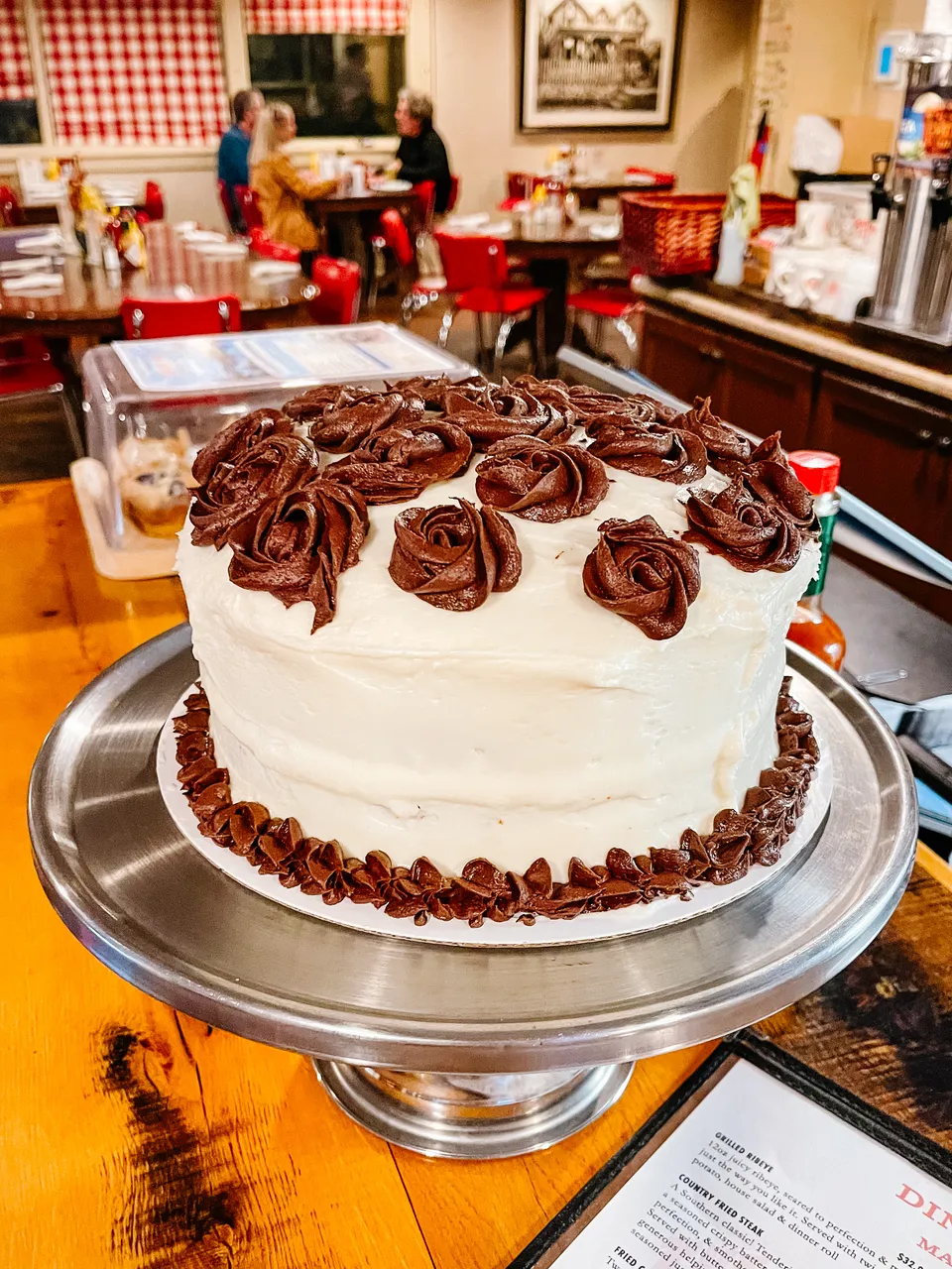 A layer cake piped with chocolate-frosting roses on a stand at the end of the counter — tonight’s case.