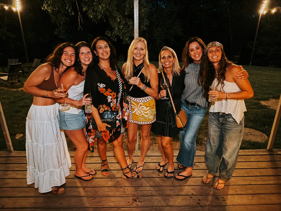 Seven friends lined up on the back deck at dusk, drinks up, the yard lit behind them.