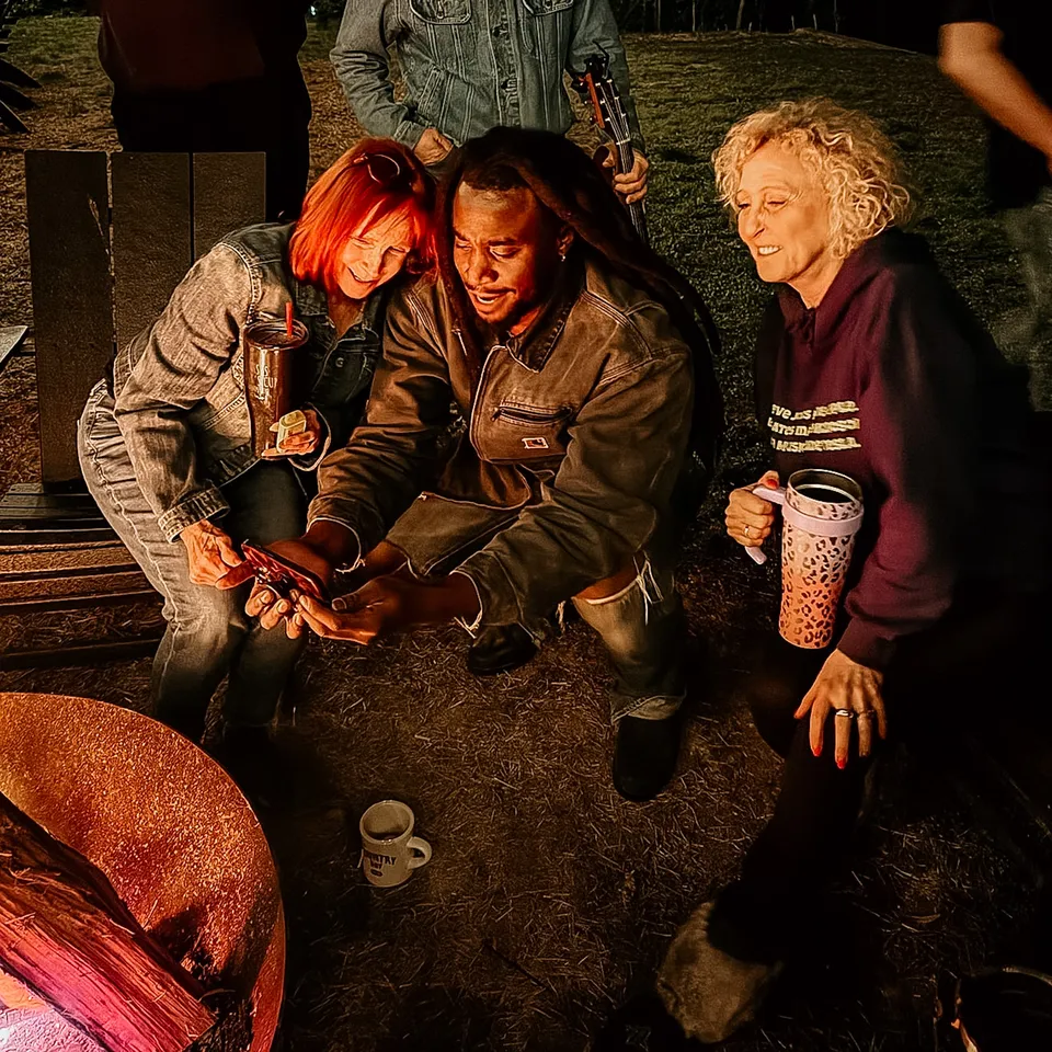 Three friends crouched at the edge of the fire, laughing at something on a phone, mugs in hand.