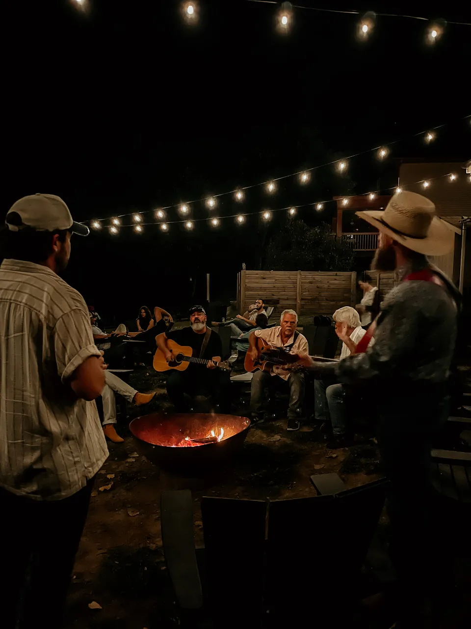Two players with acoustic guitars in the ring of chairs around the fire pit, string lights overhead.