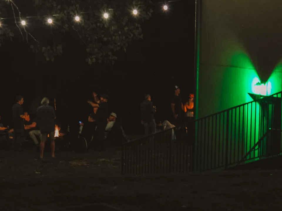 The green lamp's glow on the corner of the building at night, the stair railing below it, the fire pit crowd out in the dark beyond.