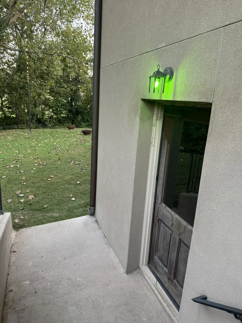 The basement door at the bottom of the outside stairs, the green lantern lit above it, the backyard beyond.