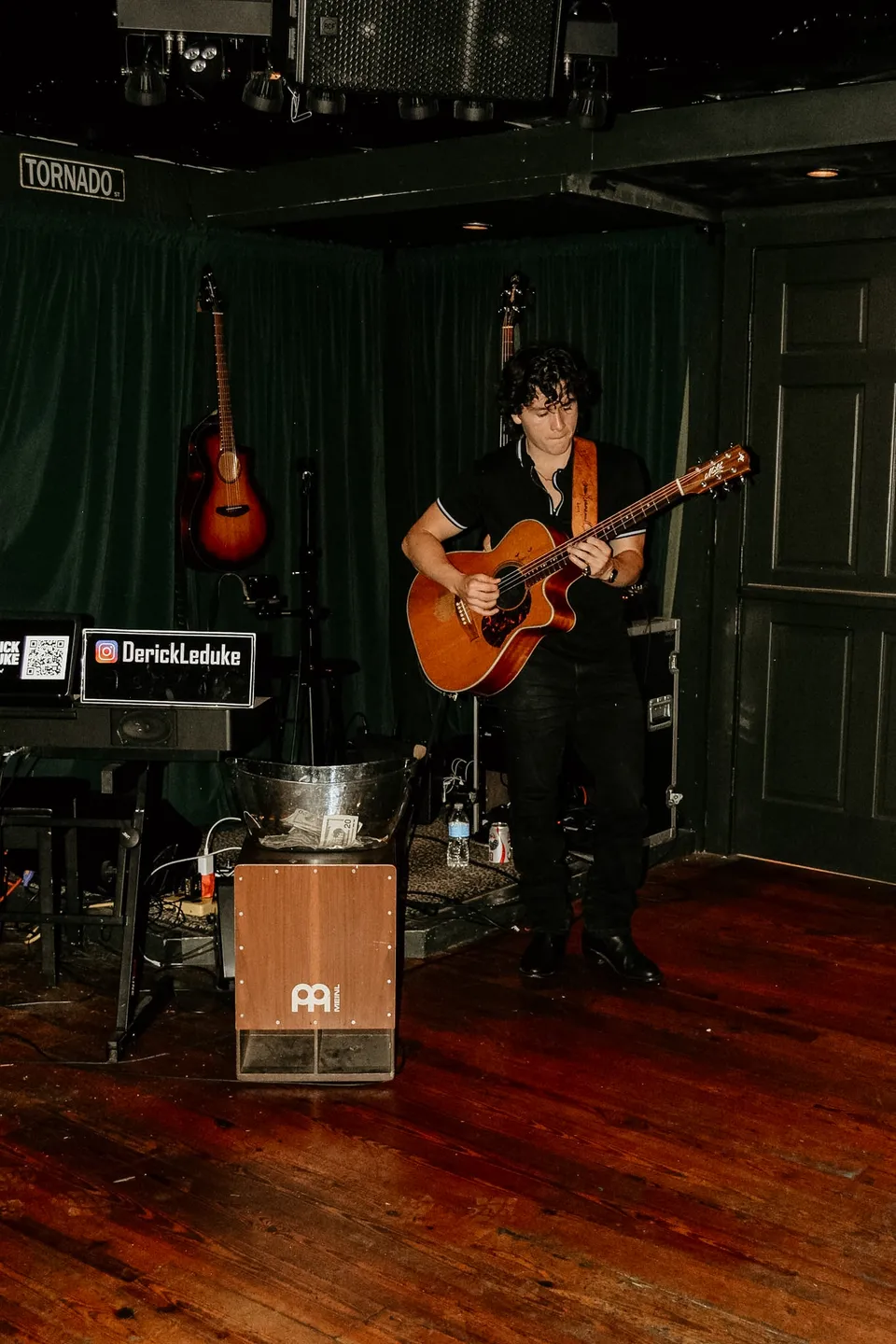 A singer with an acoustic guitar on stage, a spare guitar hung on the curtain behind, the TORNADO sign over the door.