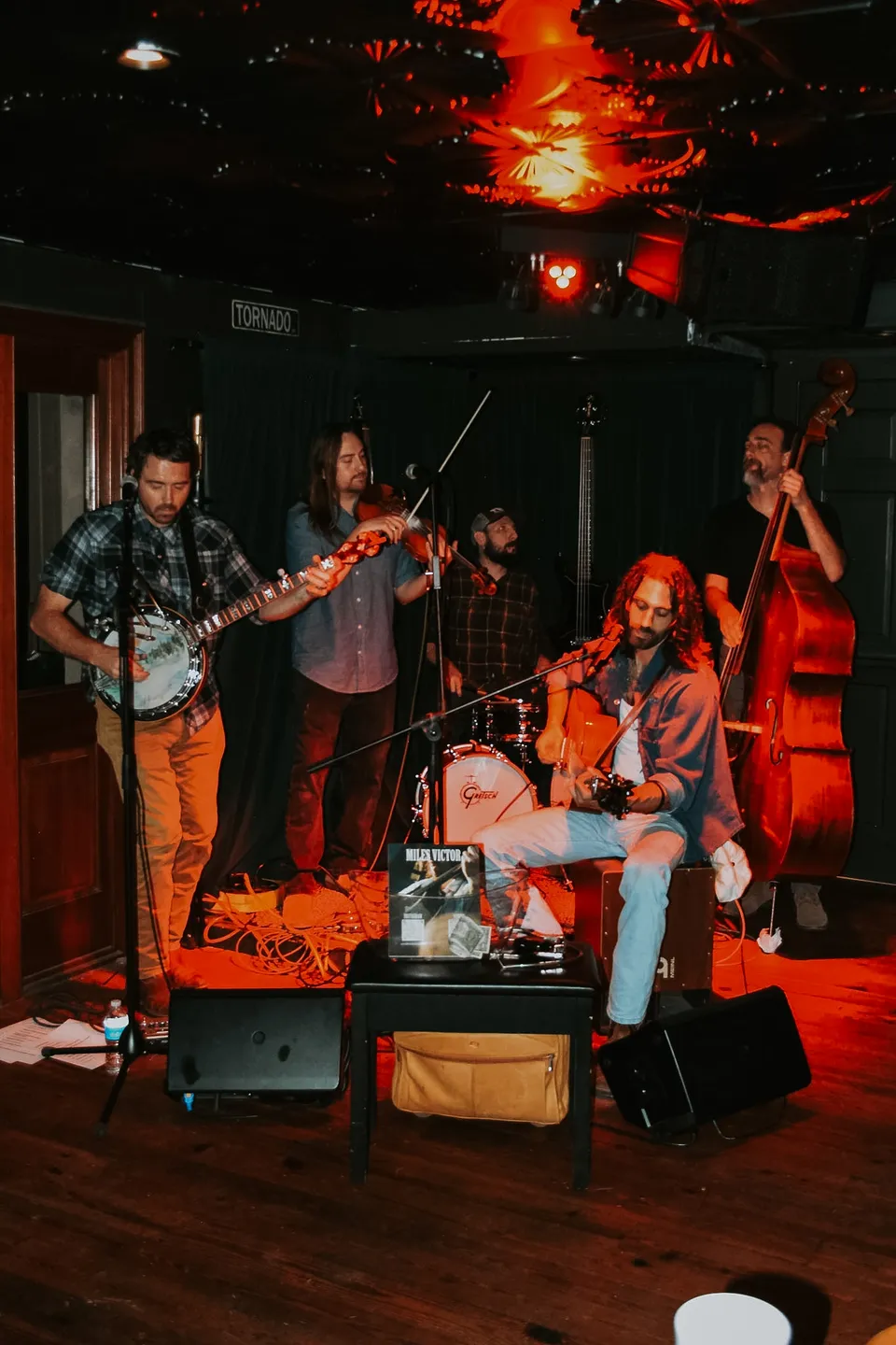 A five-piece string band — banjo, fiddle, guitar, upright bass — on the stage under red light.